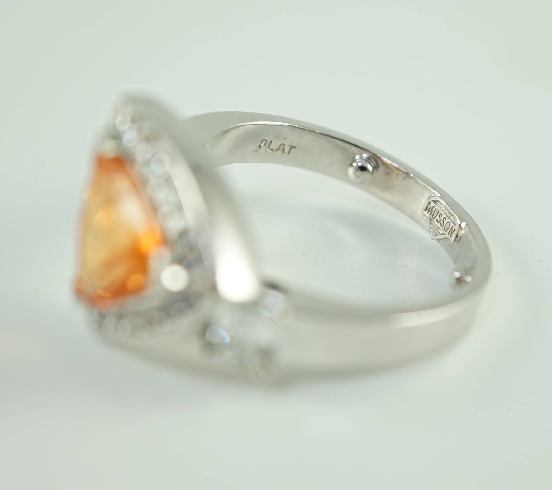 A modern platinum and single stone trilliant cut spessartite garnet ring, with diamond set border and two criss-emerald cut diamond set shoulders, by Musson